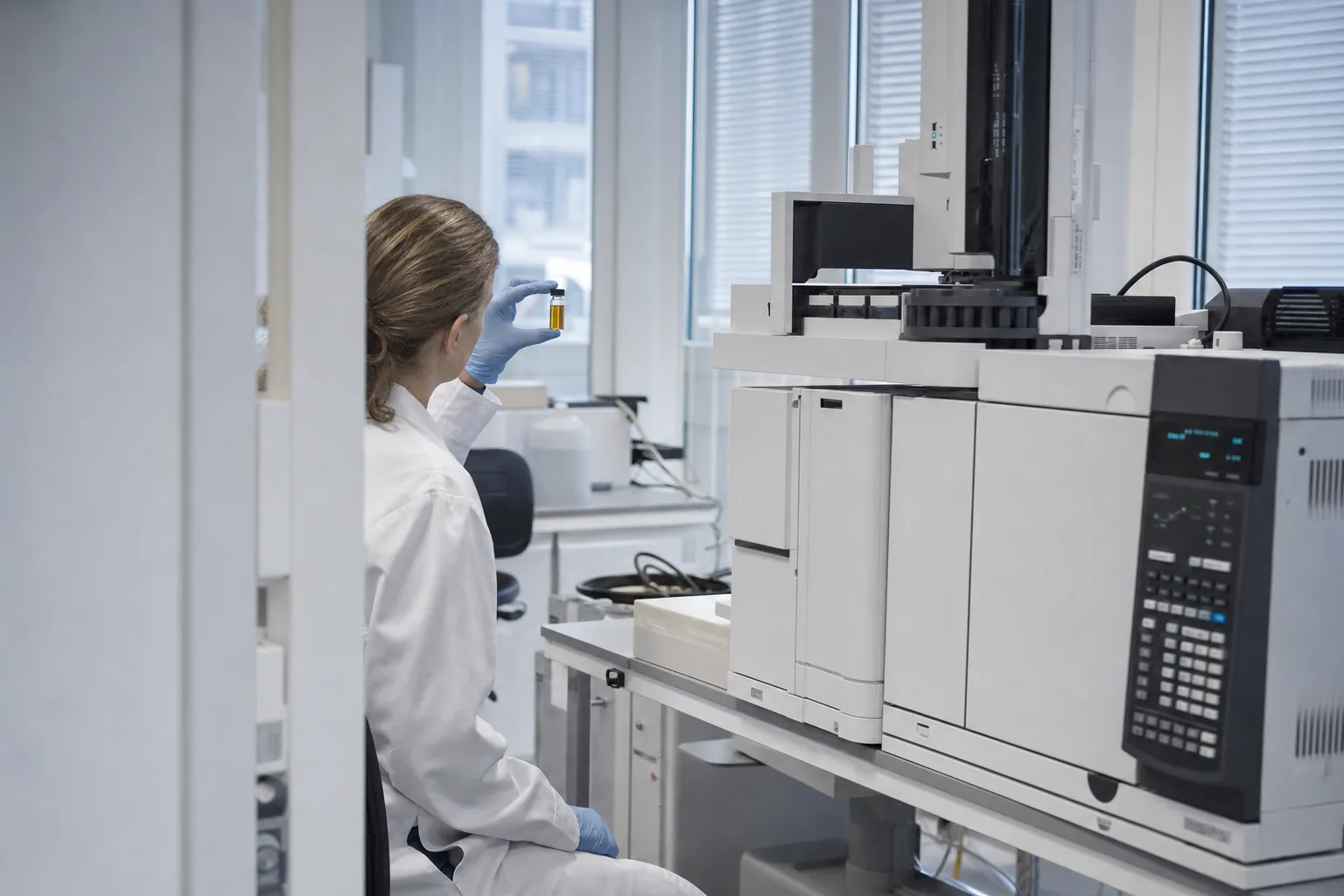 Researcher in a white lab coat at a gas chromatography instrument, inspecting a vial of amber marine oil, face turned away